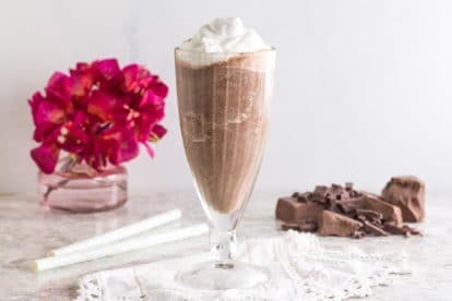 Low FODMAP Frozen Hot Chocolate in glass goblet with pink flowers, straws and chopped chocolate in background on white surface