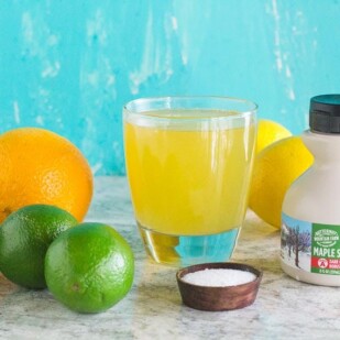 Maple Lime Low FODMAP Sports drink in a glass surrounded by ingredients against an aqua backdrop_