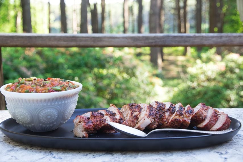 Quick Grilled Pork Tenderloin sliced on a black platter on the deck with grilled tomato peach salsa alongside