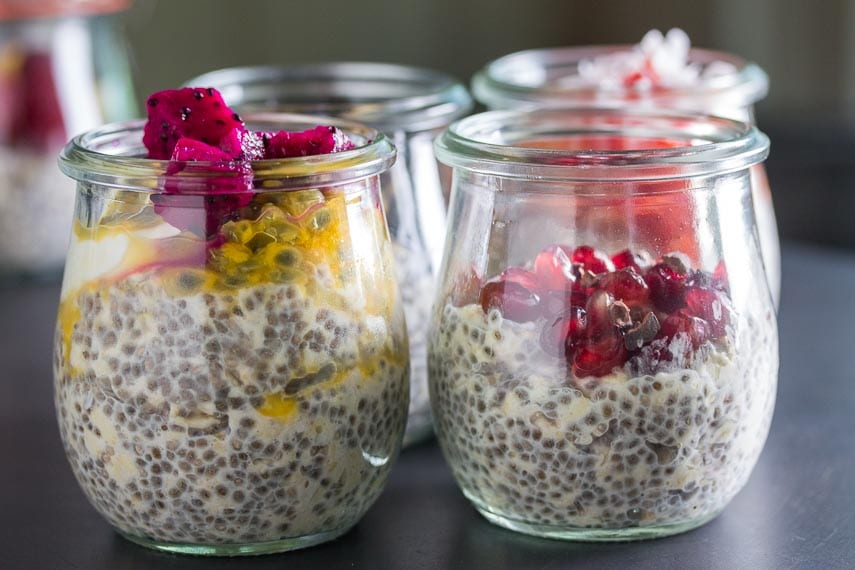 closeup of two glass jars of overnight oats and chia; one on left topped with dragon fruit and passionfruit; one on right topped with pomegranate and cacao nibs