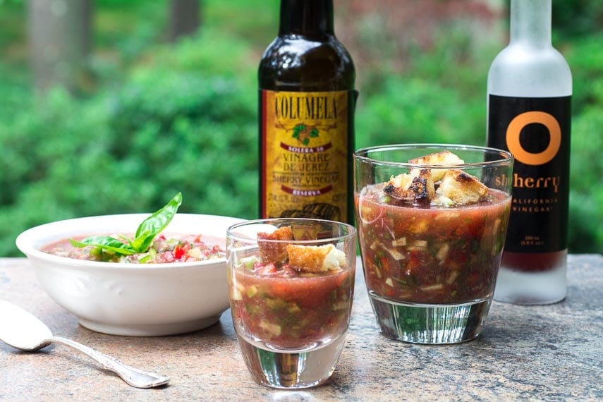 gazpacho in a bowl, garnished with basil, and two sizes of clear glasses for sipping; two kinds of sherry vinegar in background