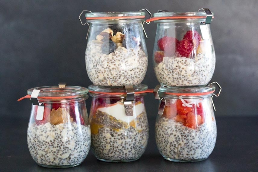 overnight oats and chia in glass jars, topped with various fruit, against black background