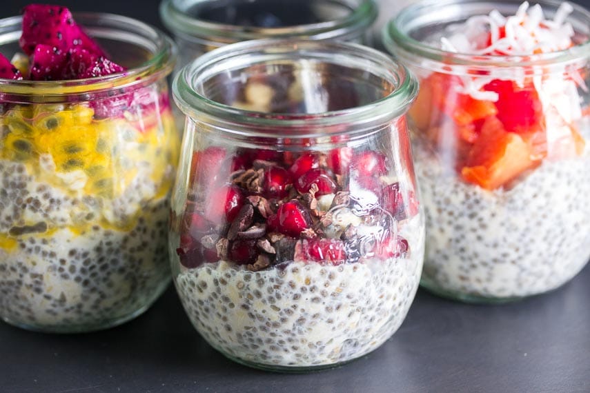trio of glass jars containing overnight oats and chia seeds, topped with different fruits; focus on pomegranate and cacao nibs