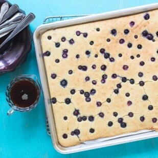 Low FODMAP Sheet Pan Pancakes with Blueberries in pan on cooling rack against aqua background; maple syrup on the side in glass pitcher