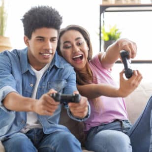 Happy black teenagers playing video game with control pad, holding joysticks in living room