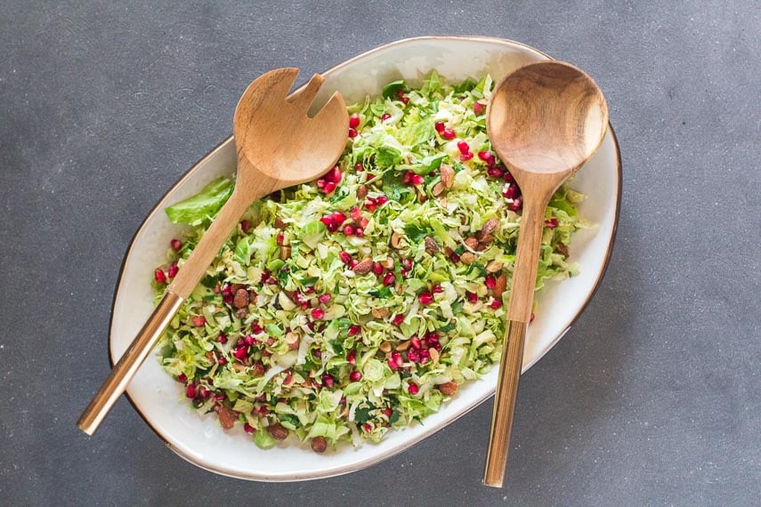 overhead image of Low FODMAP Brussels sprouts salad with pomegranate and almonds on white oval platter on dark surface