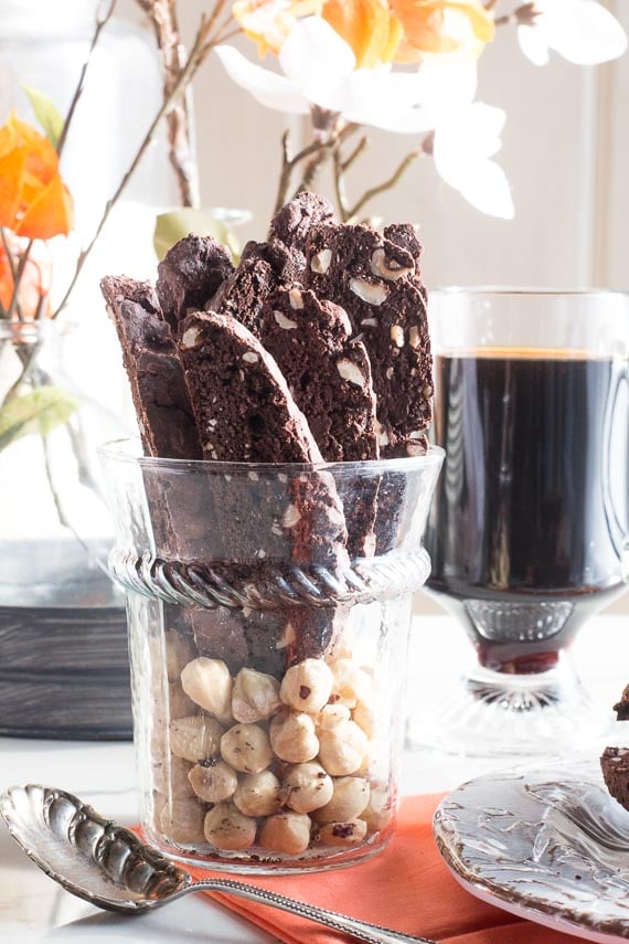 Low FODMAP Chocolate Hazelnut Biscotti in glass with decorative silver spoon in front and glass mug of coffee in the back