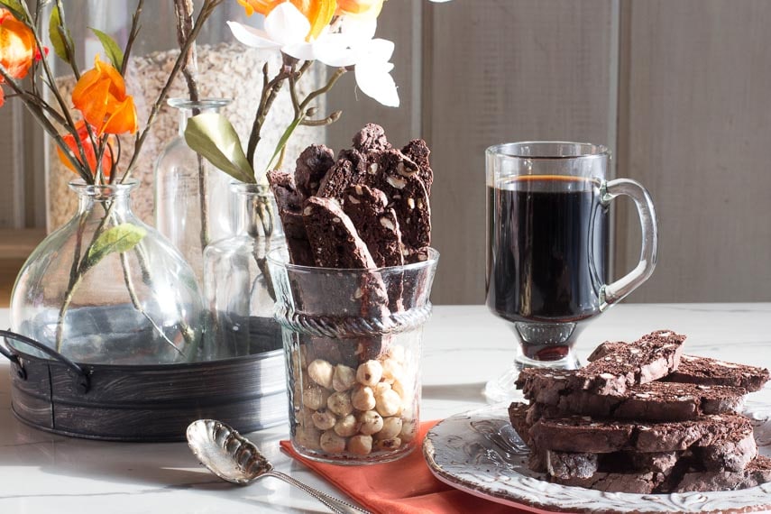 Low FODMAP Chocolate Hazelnut Biscotti stacked on a plate and upright in a glass with flowers and a mug of coffee alongside. These are decadent gluten-free Christmas cookies.