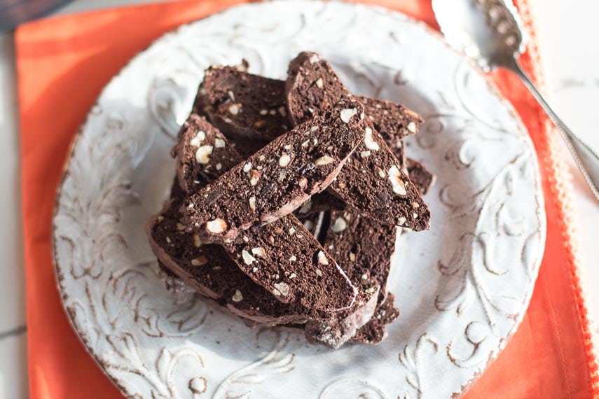 overhead image of Low FODMAP Chocolate Hazelnut Biscotti stacked on a decorative white plate, which is on top of an orange napkin
