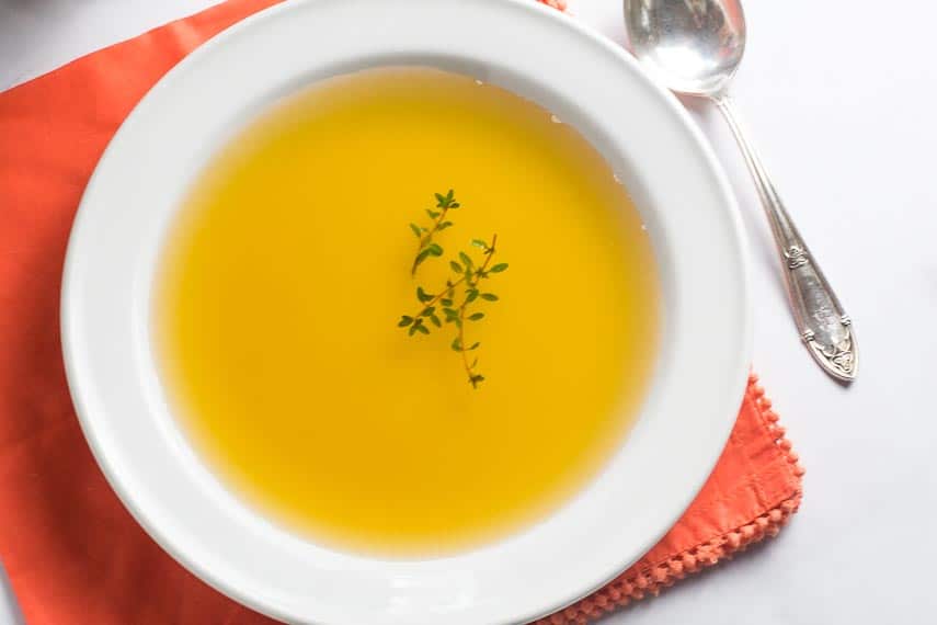 overhead image of carrot consomme in white bowl with fresh thyme floating on top; orange napkin underneath