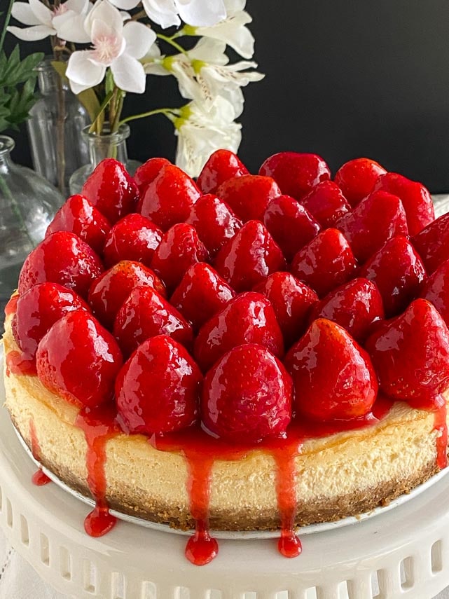 vertical pic of strawberry glazed cheesecake on white pedestal; dark background