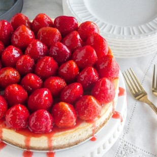 whole strawberry glazed NY style cheesecake on white pedestal with gold forks alongside