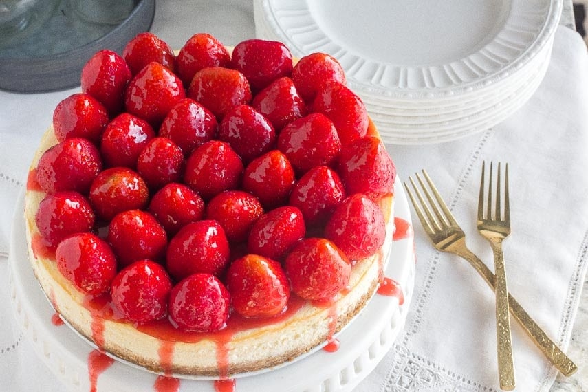 whole strawberry glazed NY style cheesecake on white pedestal with gold forks alongside