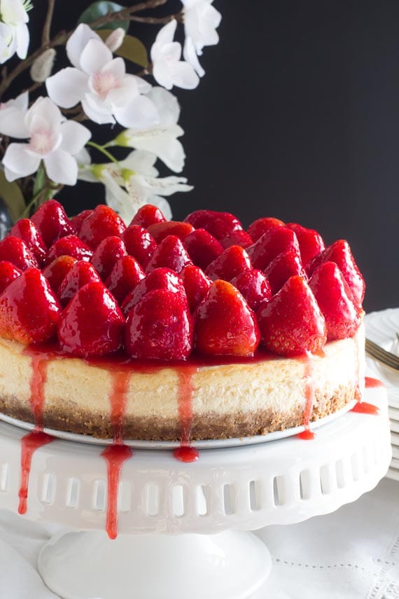 whole strawberry glazed cheesecake on white pedestal; dark background and white flowers in rear