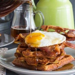 Low FODMAP Cheddar Waffles, stacked, with bacon and egg and maple syrup poured on top, all on a grey plate