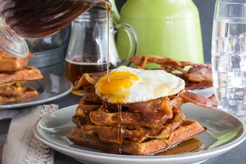 Low FODMAP Cheddar Waffles, stacked, with bacon and egg and maple syrup poured on top, all on a grey plate