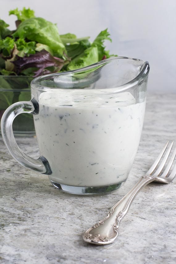 closeup vertical image of Low FODMAP Ranch Dressing in clear glass pitcher; green salad in background
