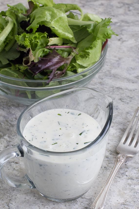 vertical image of Low FODMAP Ranch Dressing in clear glass pitcher; green salad in background