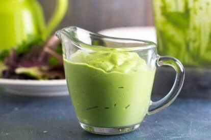 VEGAN Low FODMAP Avocado Green Goddess Dressing in clear pitcher; blender in background. Blue backdrop