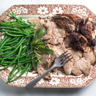 Low FODMAP Porchetta Pork Roast, sliced, with green beans on a brown and white platter with meat fork