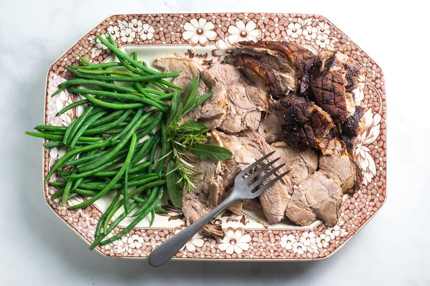 Low FODMAP Porchetta Pork Roast, sliced, with green beans on a brown and white platter with meat fork