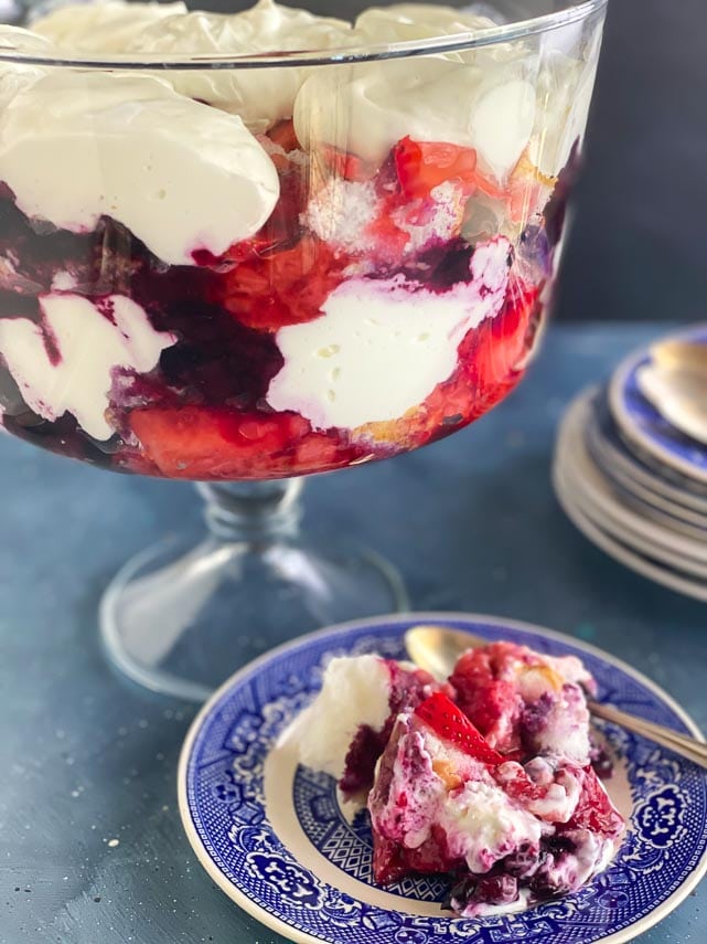 Low FODMAP Red, White & Blue Trifle in clear glass trifle bowl; individual serving on blue plate alongside with silver spoon; vertical image