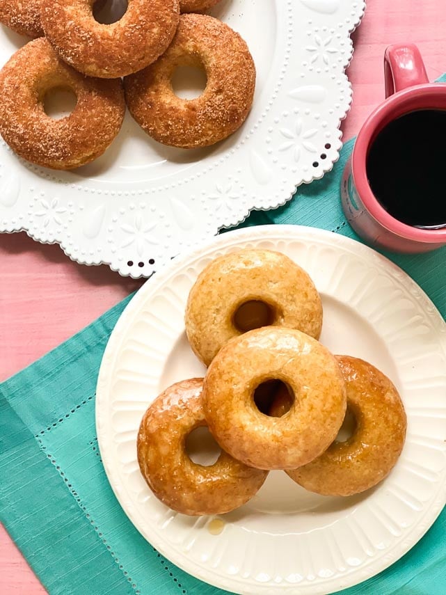 an array of low FODMAP baked doughnuts; a pile of honey glazed on a white plate and a pile of cinnamon sugar coated on a white plate. Pink mug with coffee and aqua linen napkin alongside