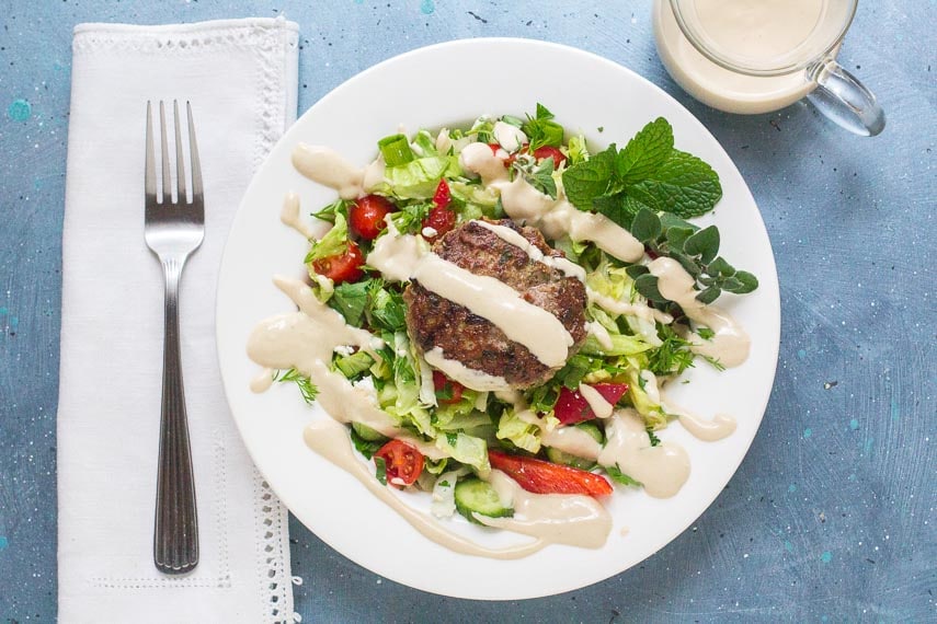 overhead shot of low FODMAP Lamb Burgers with Tahini Sauce on salad and white plate; linen napkin and fork alongside
