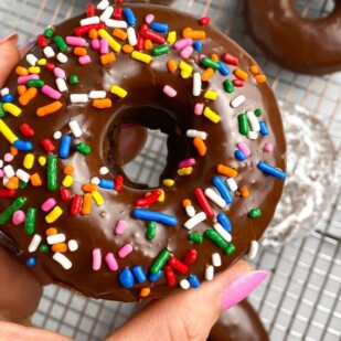 sprinkle covered low FODMAP Baked Chocolate Doughnut held in manicured hand; over rack holding doughnuts