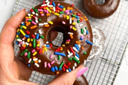 sprinkle covered low FODMAP Baked Chocolate Doughnut held in manicured hand; over rack holding doughnuts