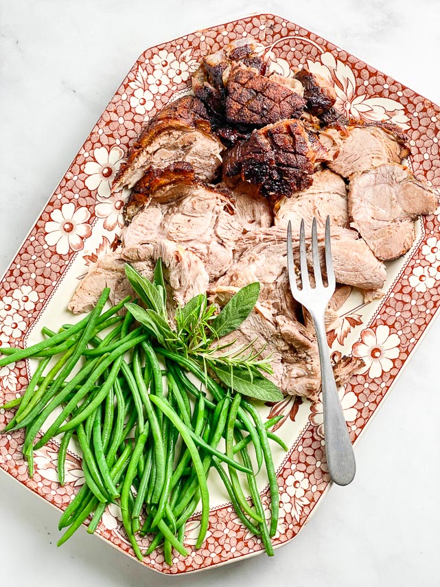 vertical image of Low FODMAP Porchetta Pork Roast on decorative brown and white platter; with green beans and meat fork