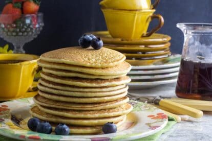 Big stack of low FODMAP high protein pancakes, on decorative plate with blueberries; small pitcher of maple syrup in background