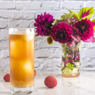 Low FODMAP Iced Black tea with Lychees in tall clear glass; white background and magenta flowers in a small vase