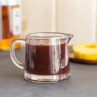 Low FODMAP Pineapply Whiskey BBQ Sauce in small glass pitcher; pineapple slices in background