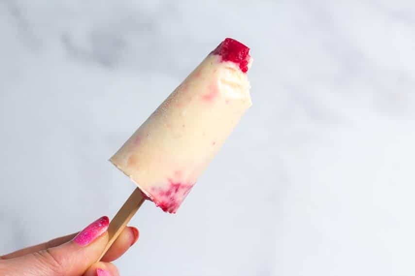 Low FODMAP peaches & cream popsicles with raspberries, on wooden stick held in manicured hand