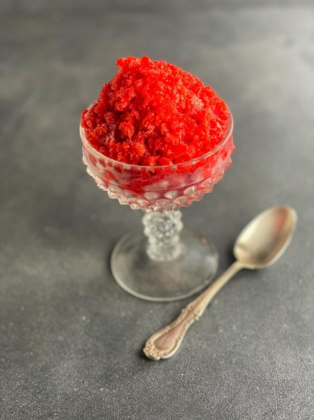 closeup of pile of strawberry granita in decorative footed glass dish, on grey surface, with silver spoon alongside