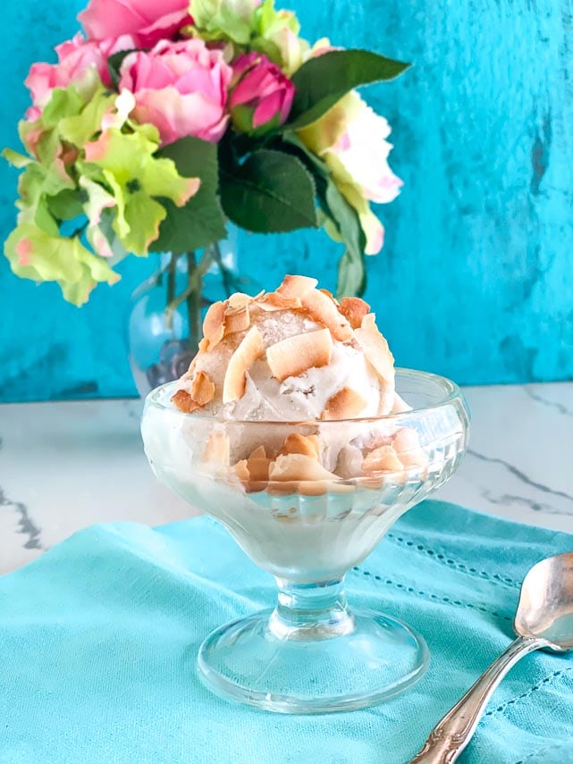 low FODMAP banana coconut sorbet with toasted coconut flakes on top in short glass dish on aqua background; silver spoon alongside