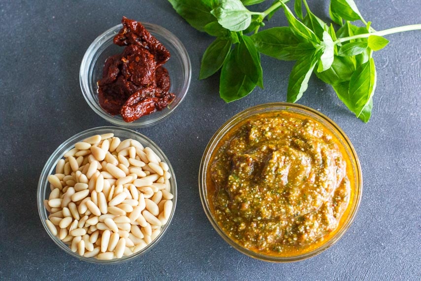 overhead view of low FODMAP sun-dried tomato pesto in glass bowl; pine nuts and basil alongside on dark surface