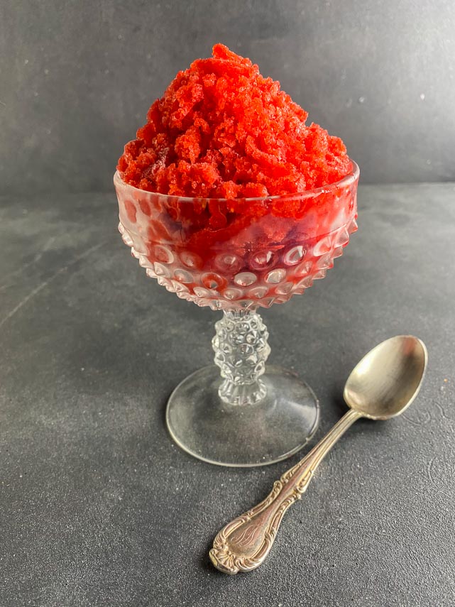 pile of strawberry granita in decorative footed glass dish, on grey surface, with silver spoon alongside