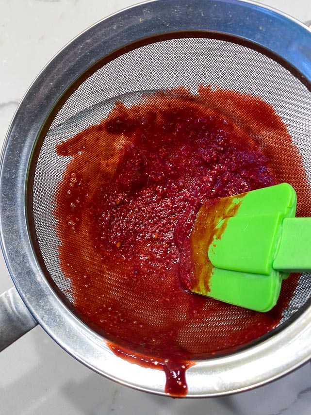 using spatula to press pureed blackberries through a strainer, leaving seeds behind