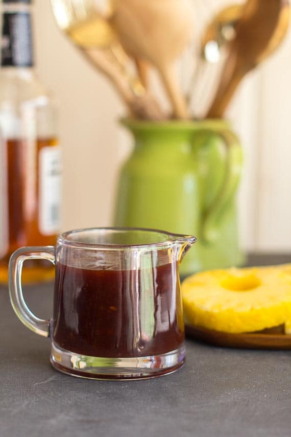 vertical image of Low FODMAP Pineapply Whiskey BBQ Sauce in small glass pitcher; pineapple slices in background