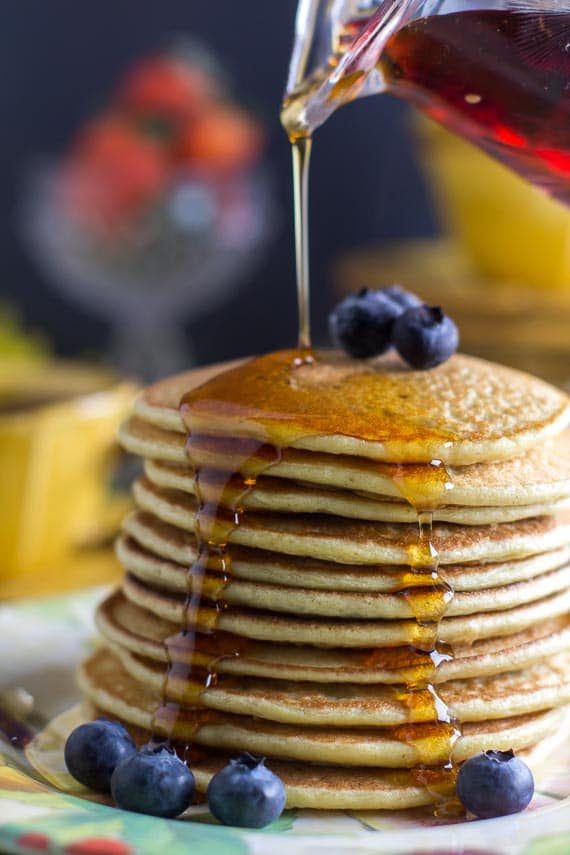 vertical image of stack of high protein pancakes on decorative plate with blueberries; syrup pouring on top