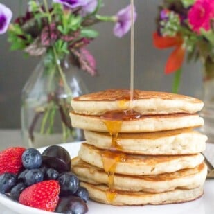 a perfect stack of low FODMAP FLUFFY Pancakes on a white plate, syrup pouring over top; fruit salad on the side