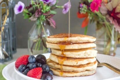 a perfect stack of low FODMAP FLUFFY Pancakes on a white plate, syrup pouring over top; fruit salad on the side