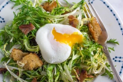 closeup of frisée salad with poached egg, bacon and sourdough croutons in white plate with blue border; silver fork alongside