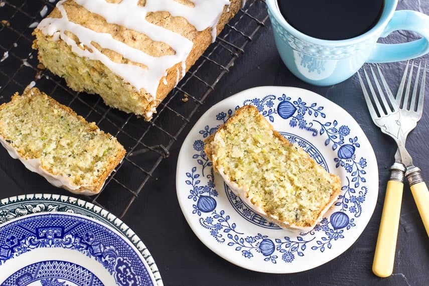 overhead image of low FODMAP Lemon Zucchini bread on decorative blue plate with aqua coffee mug; loaf is on cooling rack; antique forks alongside