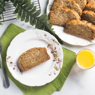 overhead of slice of zucchini banana bread on white bread; glass of orange juice alongside and fork and green napkin