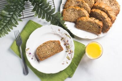 overhead of slice of zucchini banana bread on white bread; glass of orange juice alongside and fork and green napkin