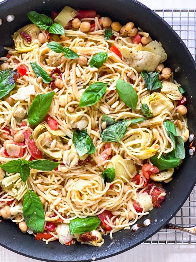 vertical image of pasta dish with basil and tomatoes in pan on cooling rack