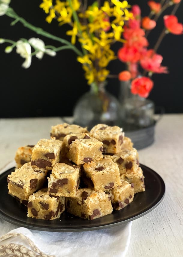 vertical image of pile of cookie dough blondies on dark plate with flowers in background